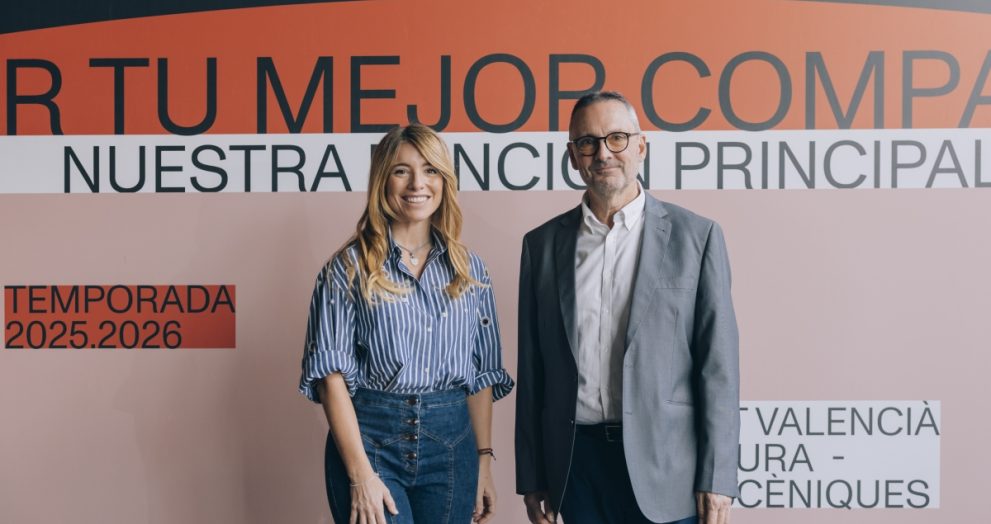 Two adults standing side by side in front of a large orange-and-pink event backdrop with Spanish text.