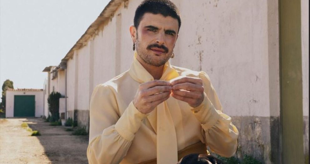 Man with a mustache wearing a pale yellow shirt ties a scarf-like accessory outdoors beside a white wall and green door in the background.