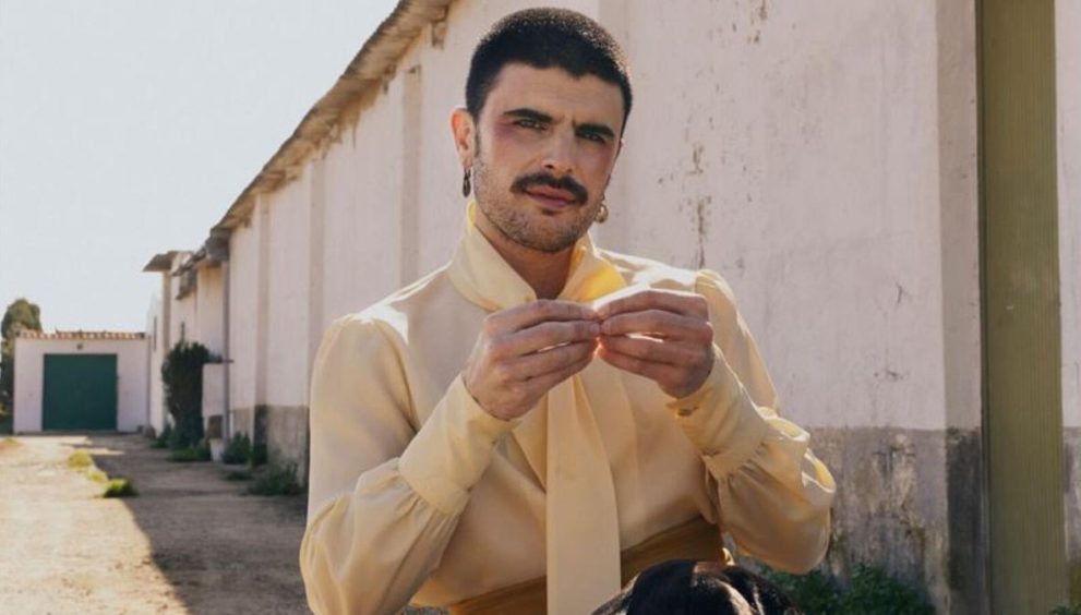 Man with a mustache wearing a pale yellow shirt ties a scarf-like accessory outdoors beside a white wall and green door in the background.
