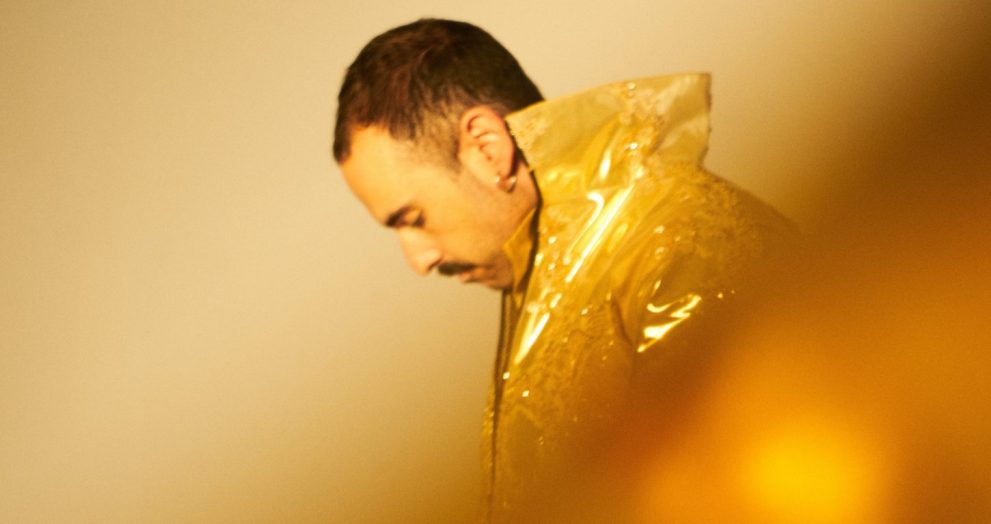 Side profile of a man with short dark hair and a mustache, wearing a shiny gold metallic cape and looking downward.