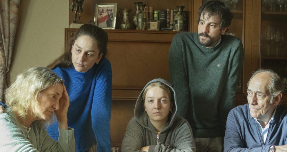 A family gathers around a table in a cozy living room, looking serious and focused on something in front of them.
