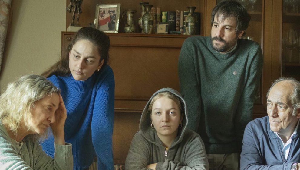 A family gathers around a table in a cozy living room, looking serious and focused on something in front of them.