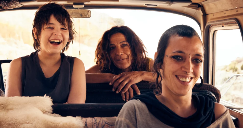 Three people riding in a van, smiling and laughing as they enjoy a sunny day together in the back seats and front passenger seat.