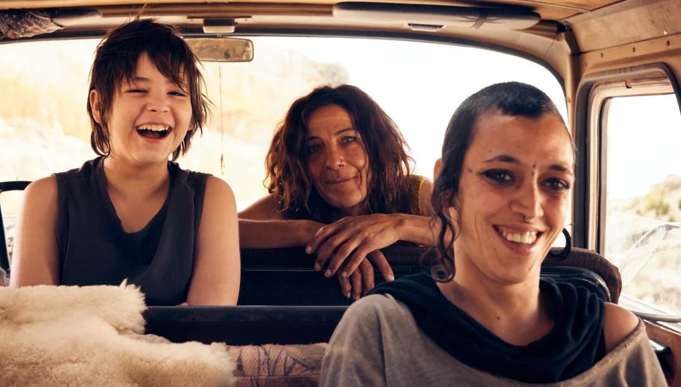 Three people riding in a van, smiling and laughing as they enjoy a sunny day together in the back seats and front passenger seat.