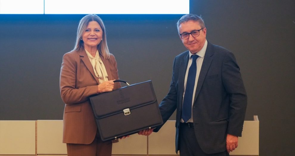 Two professionals shake hands; a woman in a brown suit holds a black briefcase, smiling at the camera in a classroom-like setting.