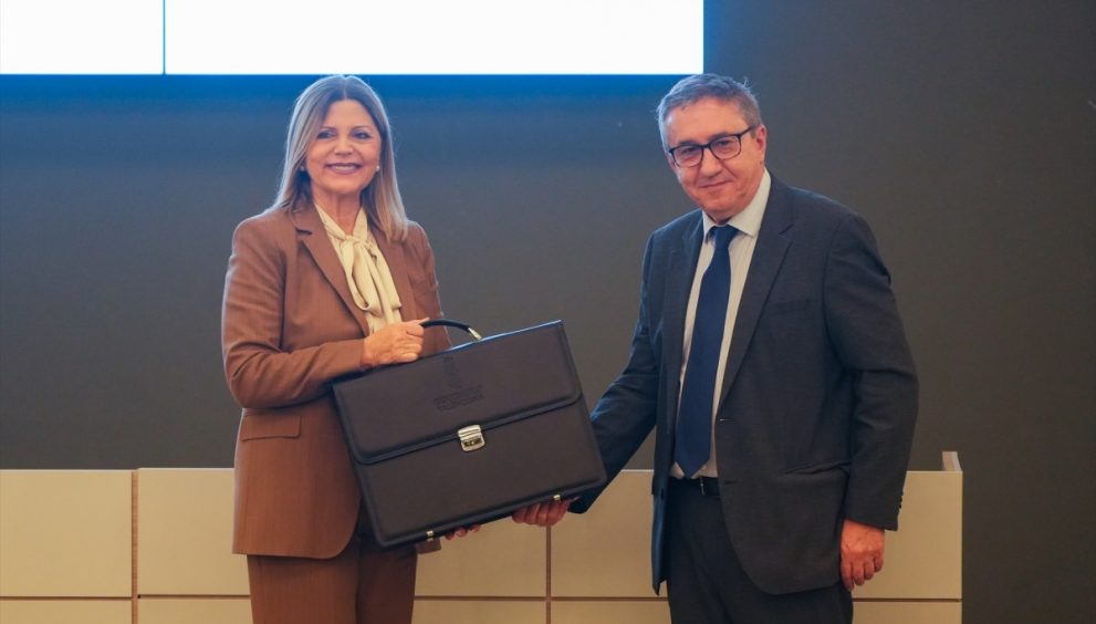 Two professionals shake hands; a woman in a brown suit holds a black briefcase, smiling at the camera in a classroom-like setting.