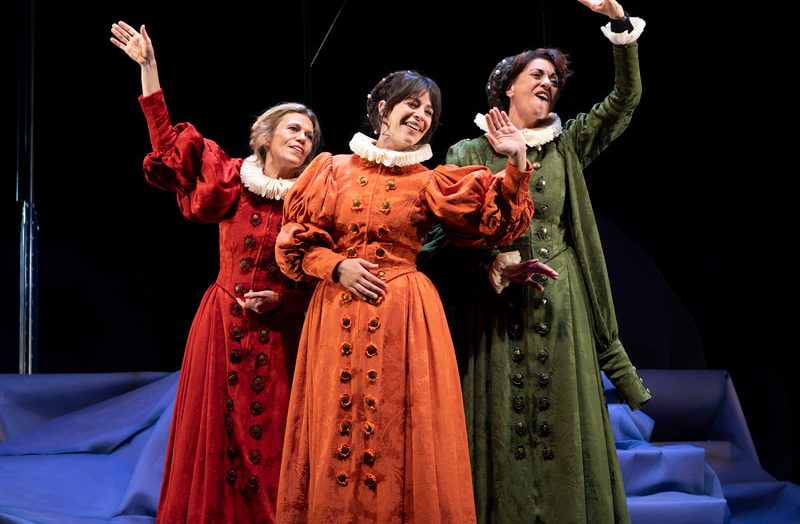 Three women in Renaissance-style dresses (red, orange, green) perform on a theater stage with raised arms.