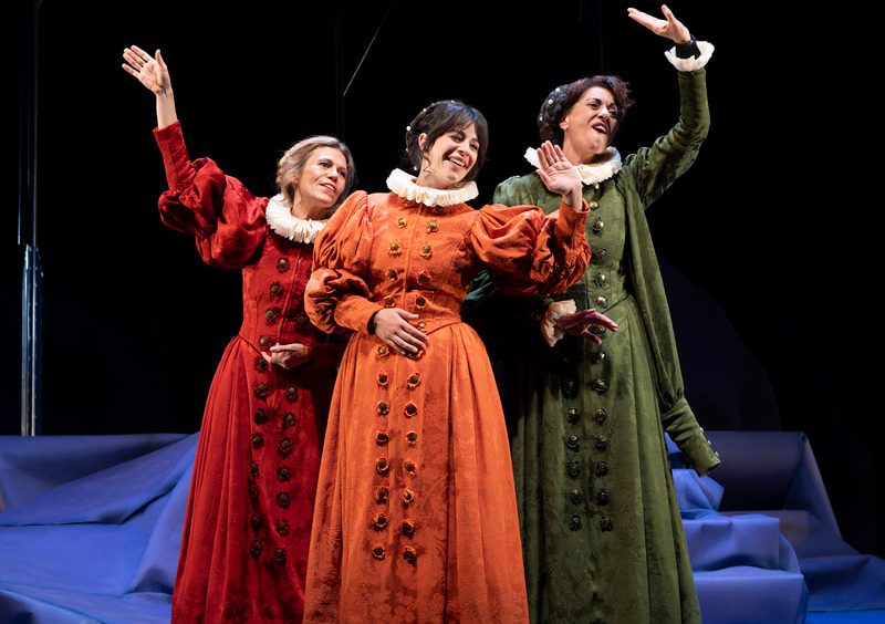 Three women in Renaissance-style dresses (red, orange, green) perform on a theater stage with raised arms.