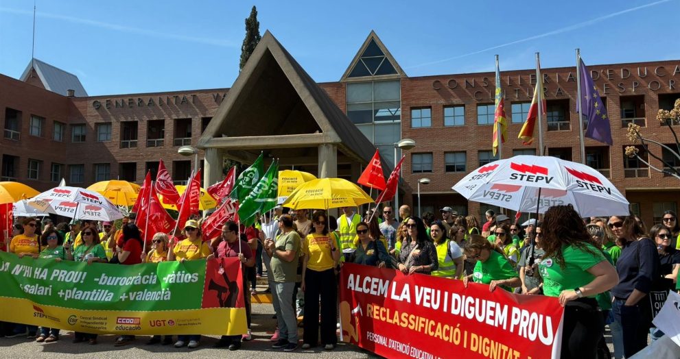 Crowd of protesters holding colorful banners and flags in front of a brick government building, wearing yellow and green shirts under a blue sky with umbrellas nearby.