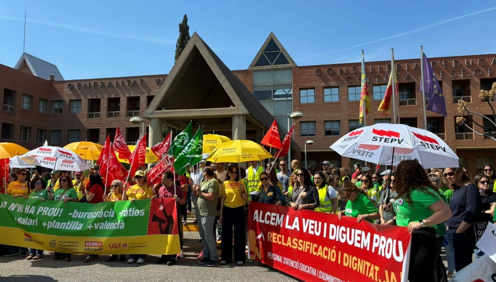 Crowd of protesters holding colorful banners and flags in front of a brick government building, wearing yellow and green shirts under a blue sky with umbrellas nearby.