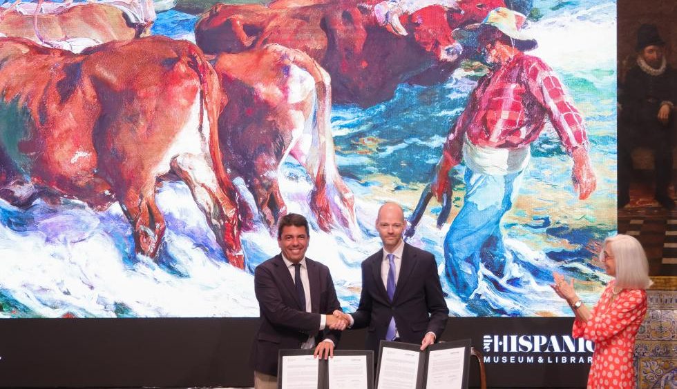 Two men in suits shake hands while signing documents at a table, with a large colorful cow mural behind them and a woman clapping nearby.