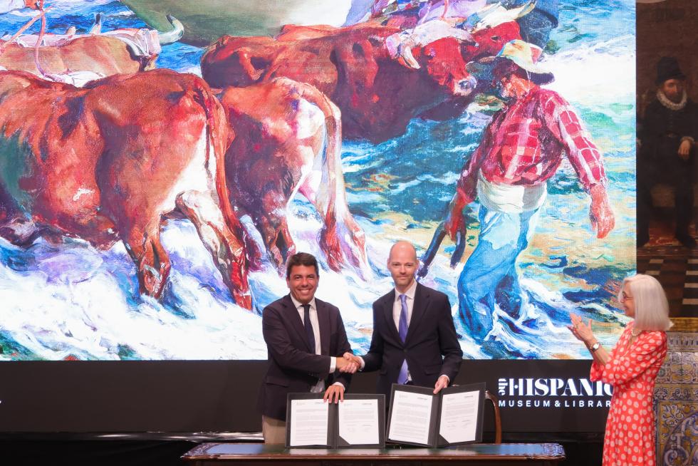 Two men in suits shake hands while signing documents at a table, with a large colorful cow mural behind them and a woman clapping nearby.