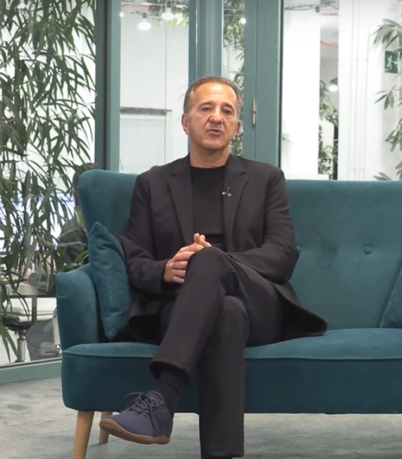 Middle-aged man in a black suit sits cross-legged on a teal sofa in a plant-filled office, wearing a lapel mic.