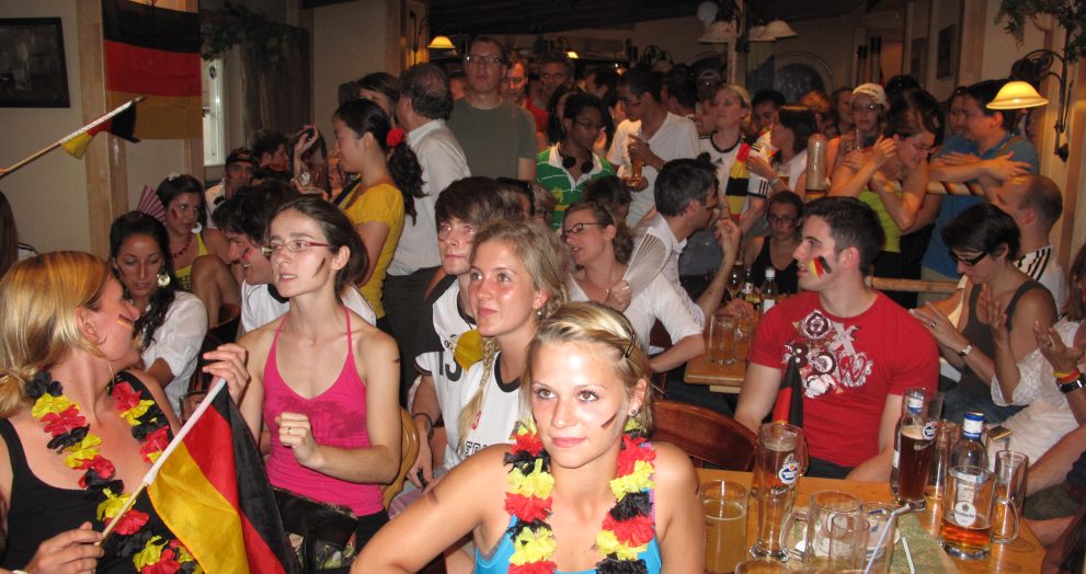 Crowded indoor party scene with people wearing colorful leis and German flags, posing and mingling around long tables with drinks and beers visible in the foreground.