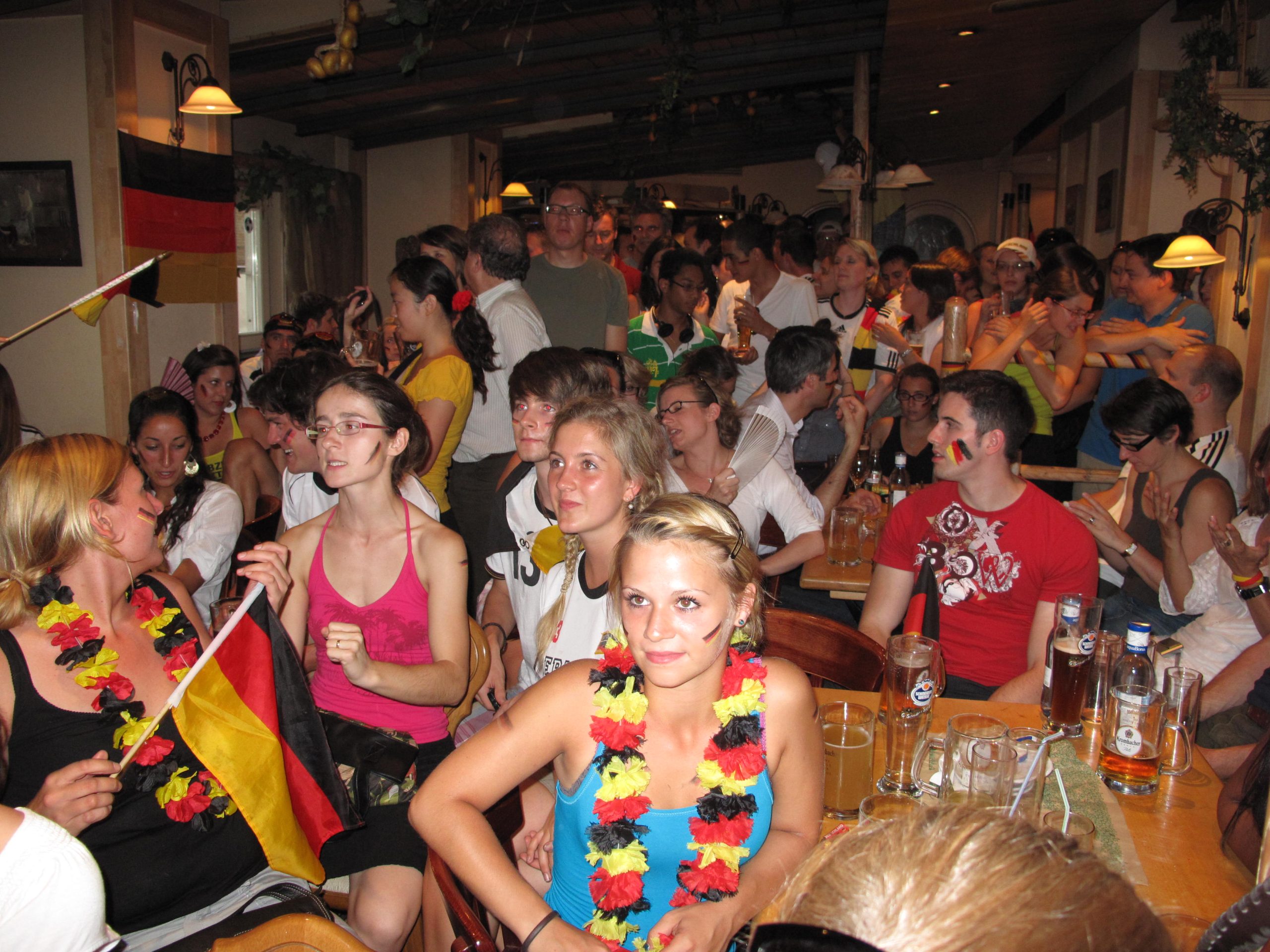 Crowded indoor party scene with people wearing colorful leis and German flags, posing and mingling around long tables with drinks and beers visible in the foreground.