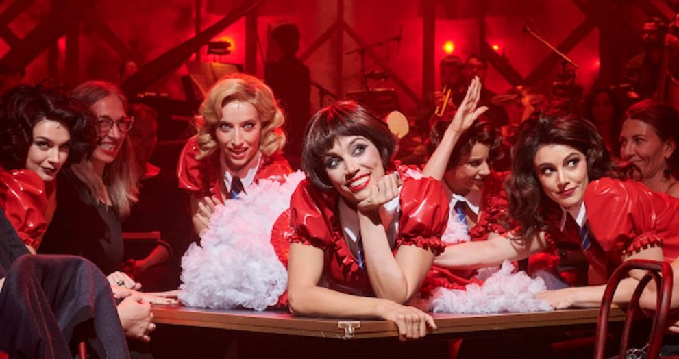 Group of performers in red costumes leaning over a table, posing and smiling during a backstage or stage show with a red-lit crowd behind them.