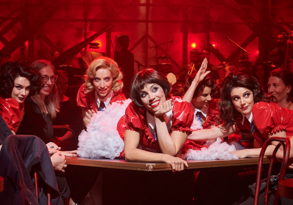 Group of performers in red costumes leaning over a table, posing and smiling during a backstage or stage show with a red-lit crowd behind them.