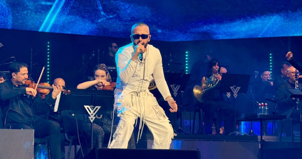 Male singer in white outfit performs center stage with a microphone while an orchestra accompanies him, blue stage lighting in the background