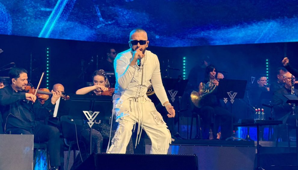 Male singer in white outfit performs center stage with a microphone while an orchestra accompanies him, blue stage lighting in the background