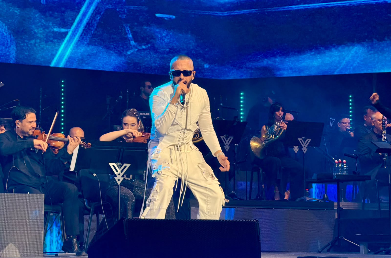 Male singer in white outfit performs center stage with a microphone while an orchestra accompanies him, blue stage lighting in the background