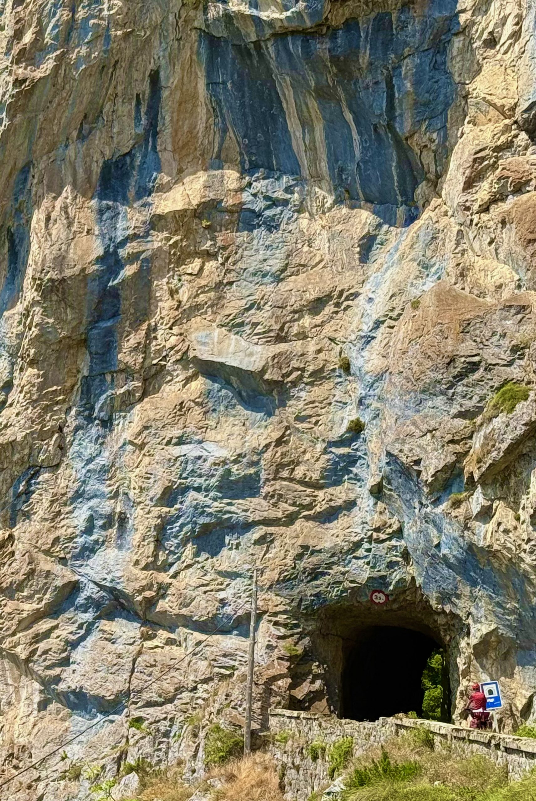 Rocky cliff with a dark tunnel entrance and a railing where two people stand nearby.