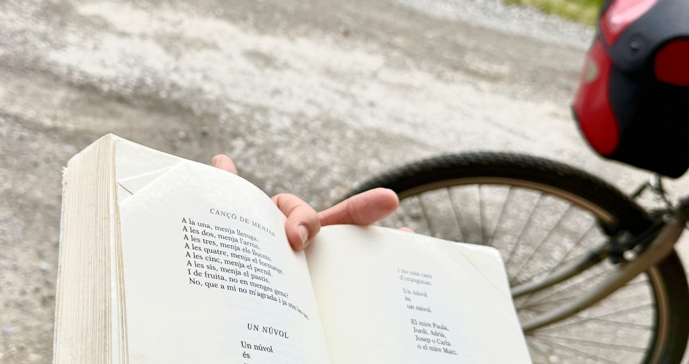 Person reads an open book on a bicycle or bike pannier, with a group on a bench and green hills in the background.