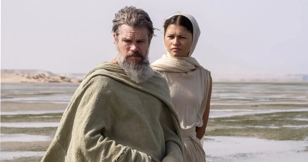 A bearded man in a beige cloak and a woman in a light headscarf stand on a flat shoreline with shallow water and distant cliffs behind them.