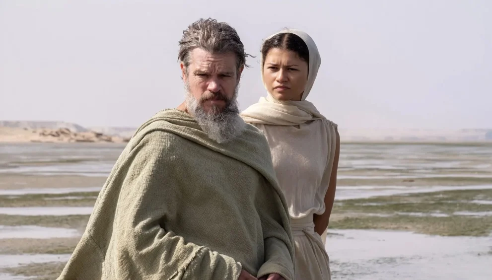 A bearded man in a beige cloak and a woman in a light headscarf stand on a flat shoreline with shallow water and distant cliffs behind them.