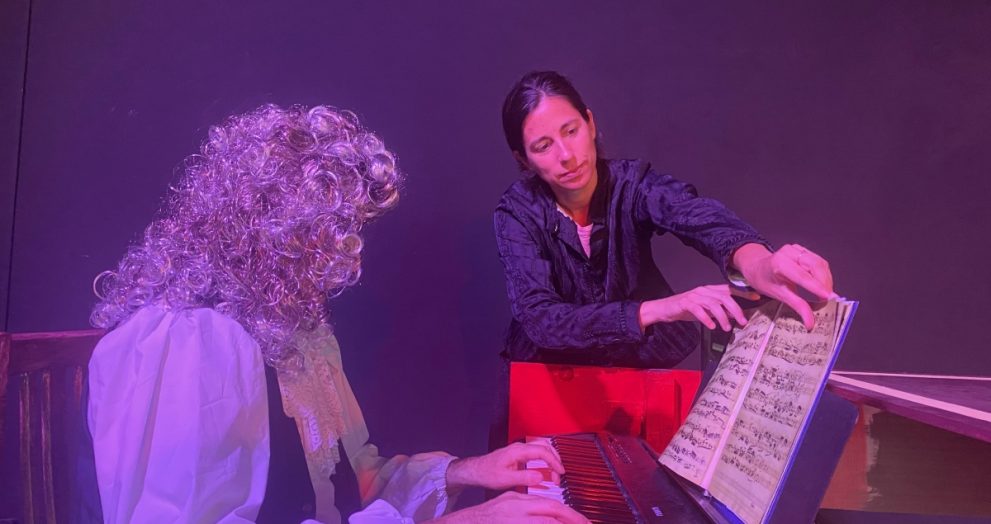 Two performers on a dimly lit stage: a curly-haired pianist in a white blouse playing a keyboard while a second person points to sheet music on a stand nearby under purple lighting.
