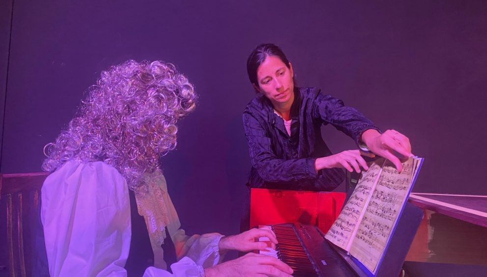 Two performers on a dimly lit stage: a curly-haired pianist in a white blouse playing a keyboard while a second person points to sheet music on a stand nearby under purple lighting.